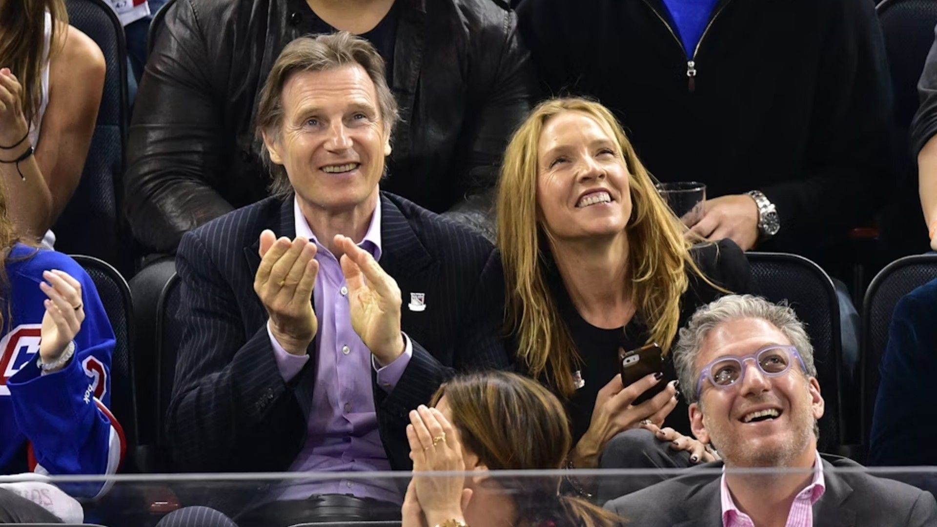 Liam Neeson y Freya St. Johnston sentados en las gradas durante un partido deportivo y animando con alegría