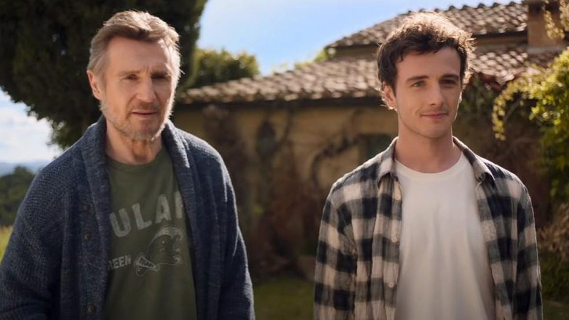 Liam Neeson y Michael Richardson están de pie uno al lado del otro frente a una casa con techo de tejas en un día soleado