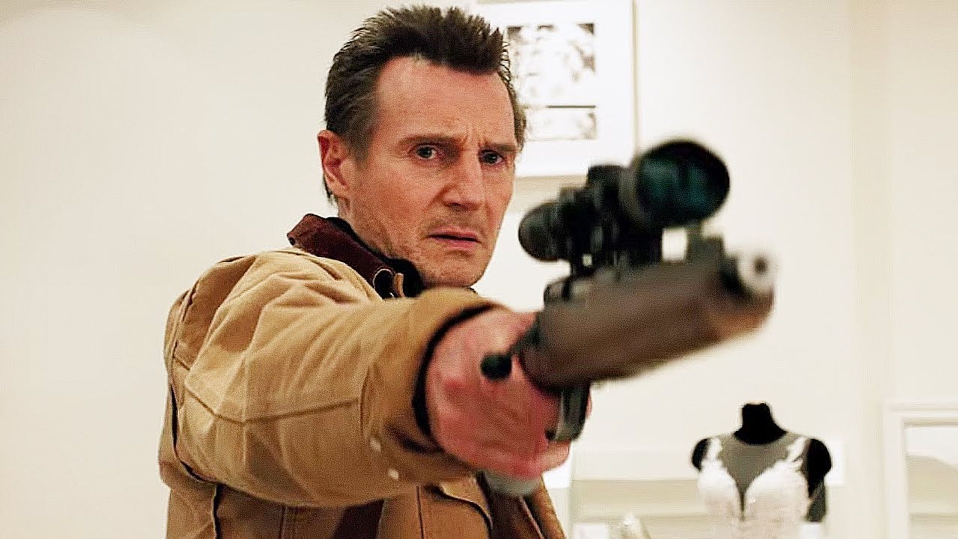 Liam Neeson en una chaqueta beige apunta un rifle con mira telescópica, con un maniquí en vestido de novia detrás de él