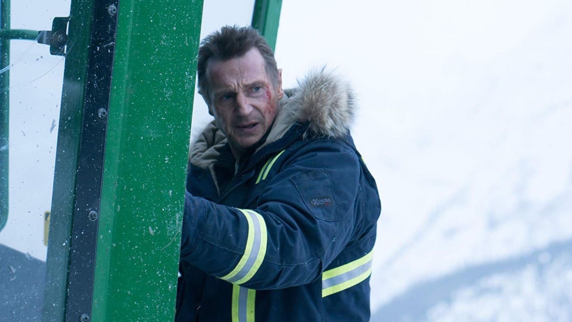 Liam Neeson con una chaqueta de invierno con capucha agarra la puerta de una cabaña verde con un paisaje cubierto de nieve al fondo