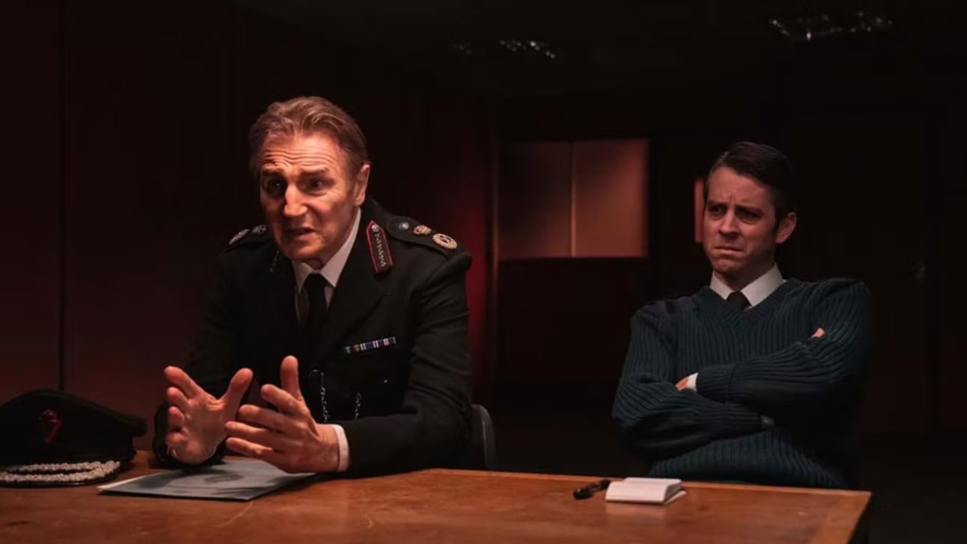 Liam Neeson con uniforme policial sentado en una mesa de interrogatorio, escena de la serie "Derry Girls"