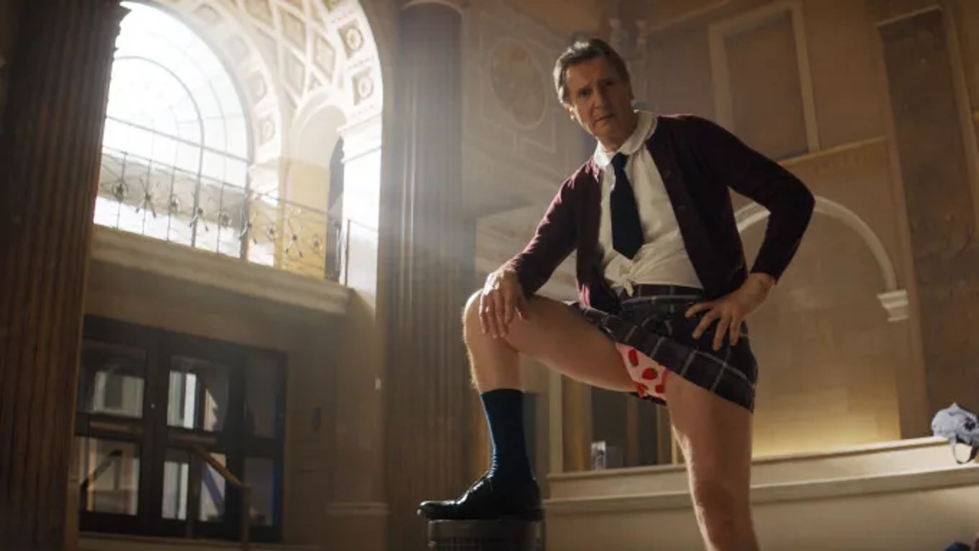 Liam Neeson con uniforme escolar y corbata posa con la pierna levantada, mostrando ropa interior con estampado de corazones, contra un fondo de columnas interiores