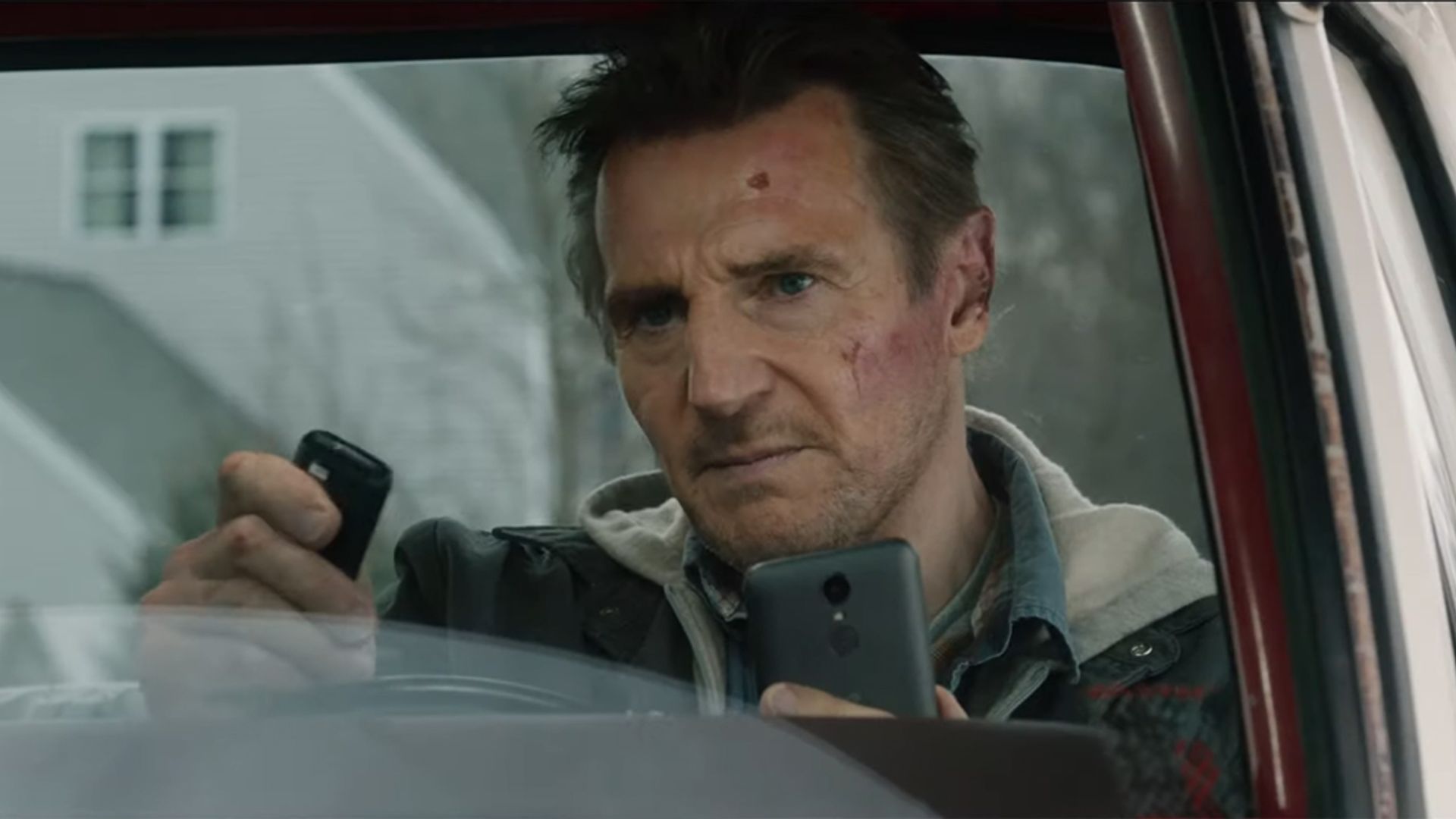 Liam Neeson está sentado en un auto mirando tensamente el teléfono y el control remoto en sus manos