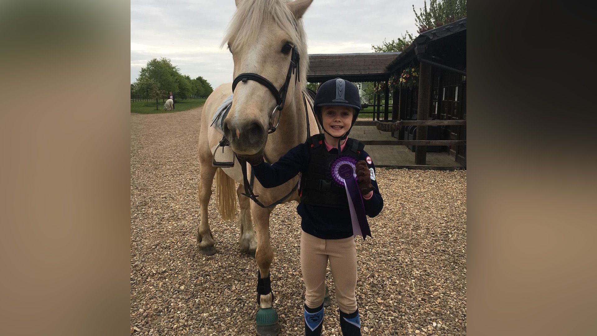 Evie Templeton de niña, vestida con ropa de equitación y casco, de pie junto a un caballo mientras sostiene una cinta de premio morada