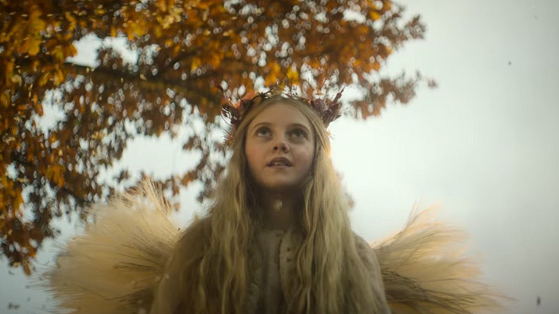 Evie Templeton con alas hechas de heno y llevando una corona de ramitas contra un fondo de cielo y árboles otoñales