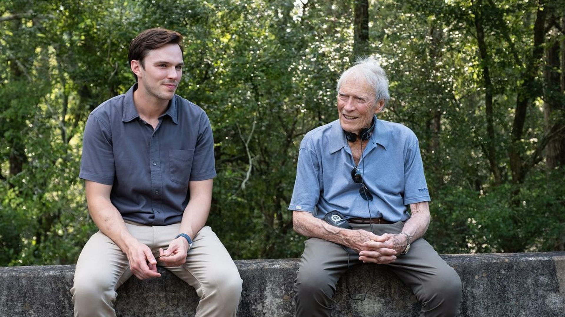 Nicholas Hoult y Clint Eastwood con ropa casual sentados en una barrera de concreto conversando con un bosque de fondo