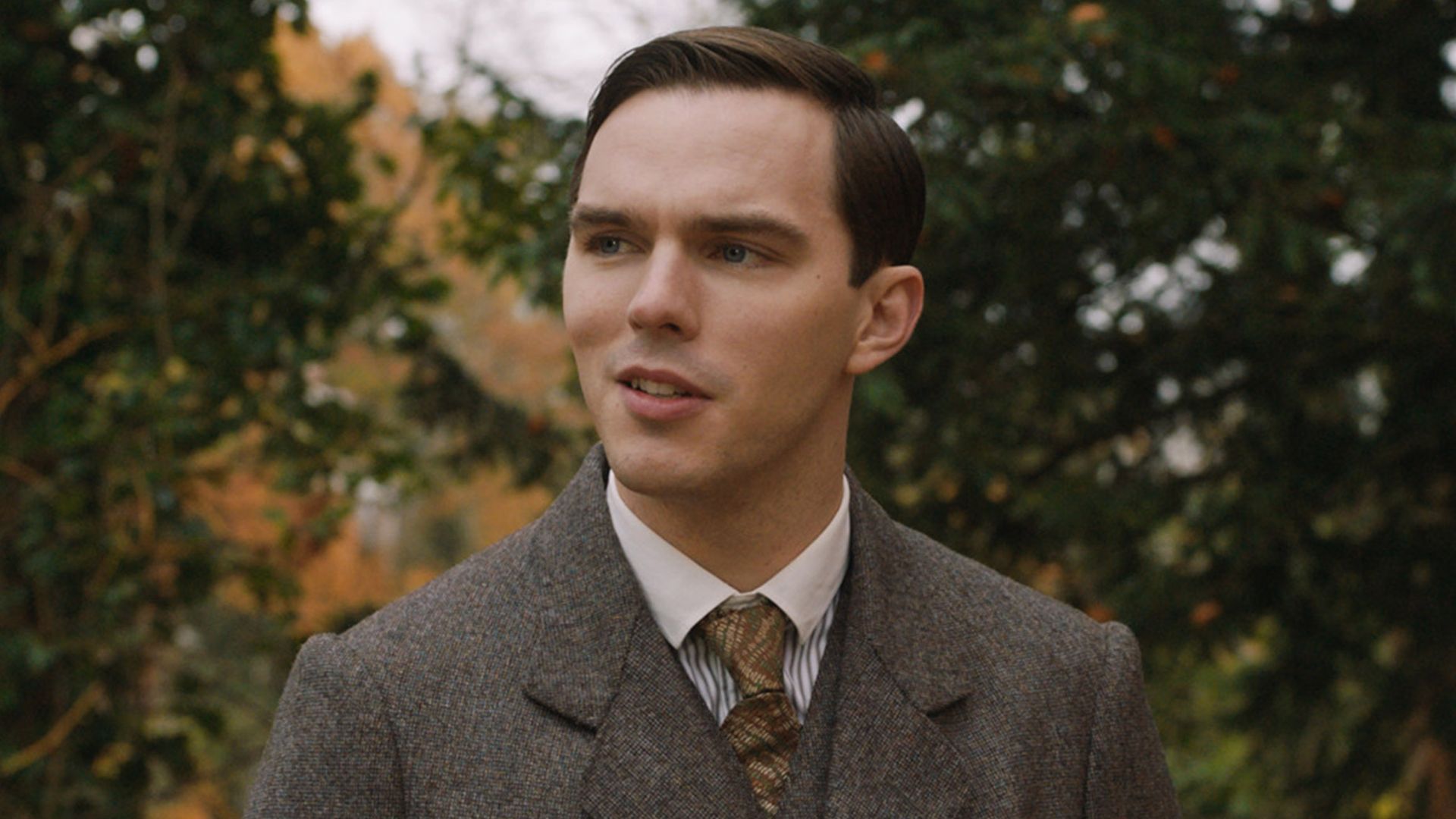 Nicholas Hoult con traje marrón y corbata de pie en la calle en el papel del joven J.R.R. Tolkien