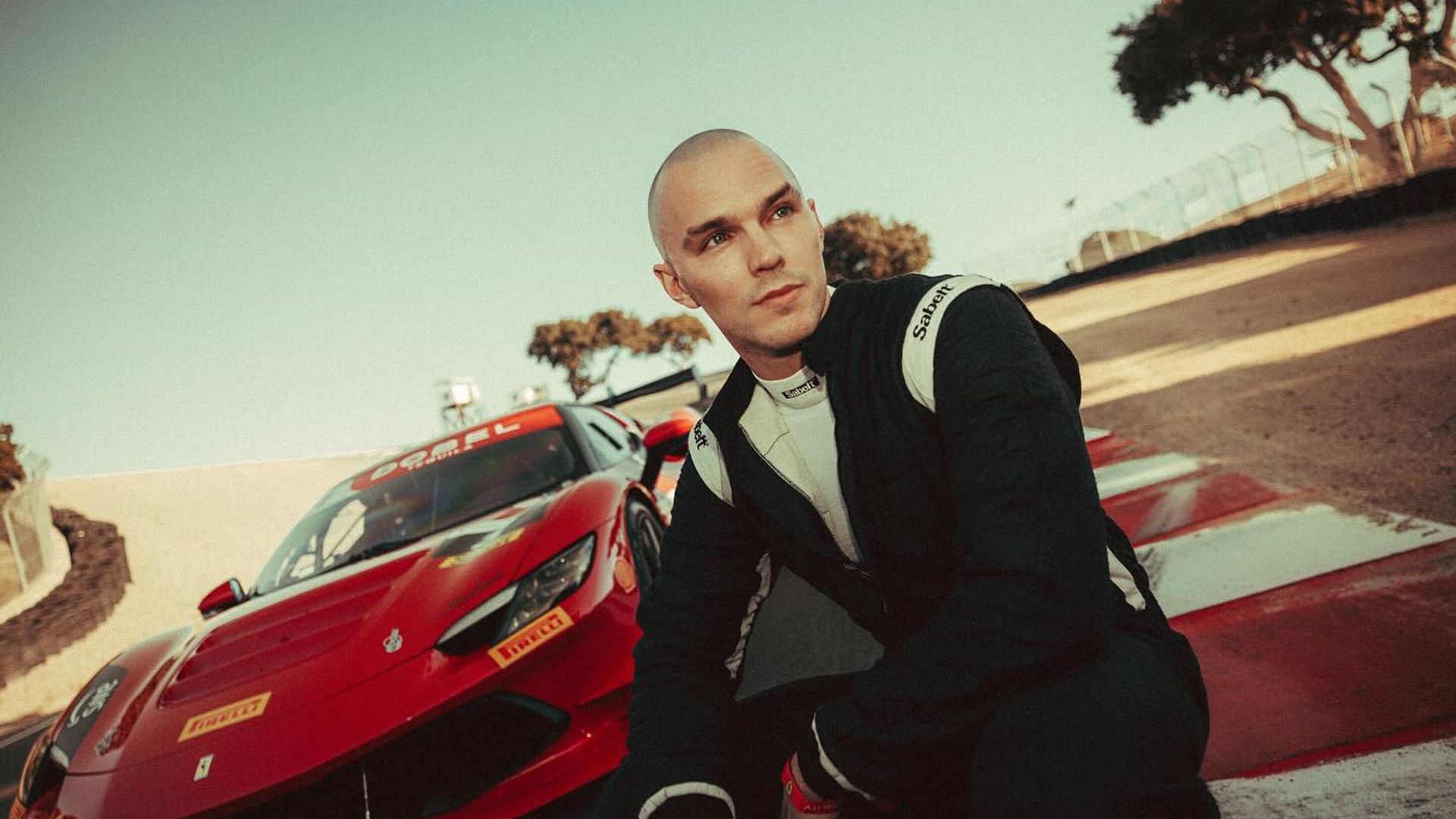 Nicholas Hoult en traje de carreras sentado en la pista con un deportivo Ferrari rojo detrás de él