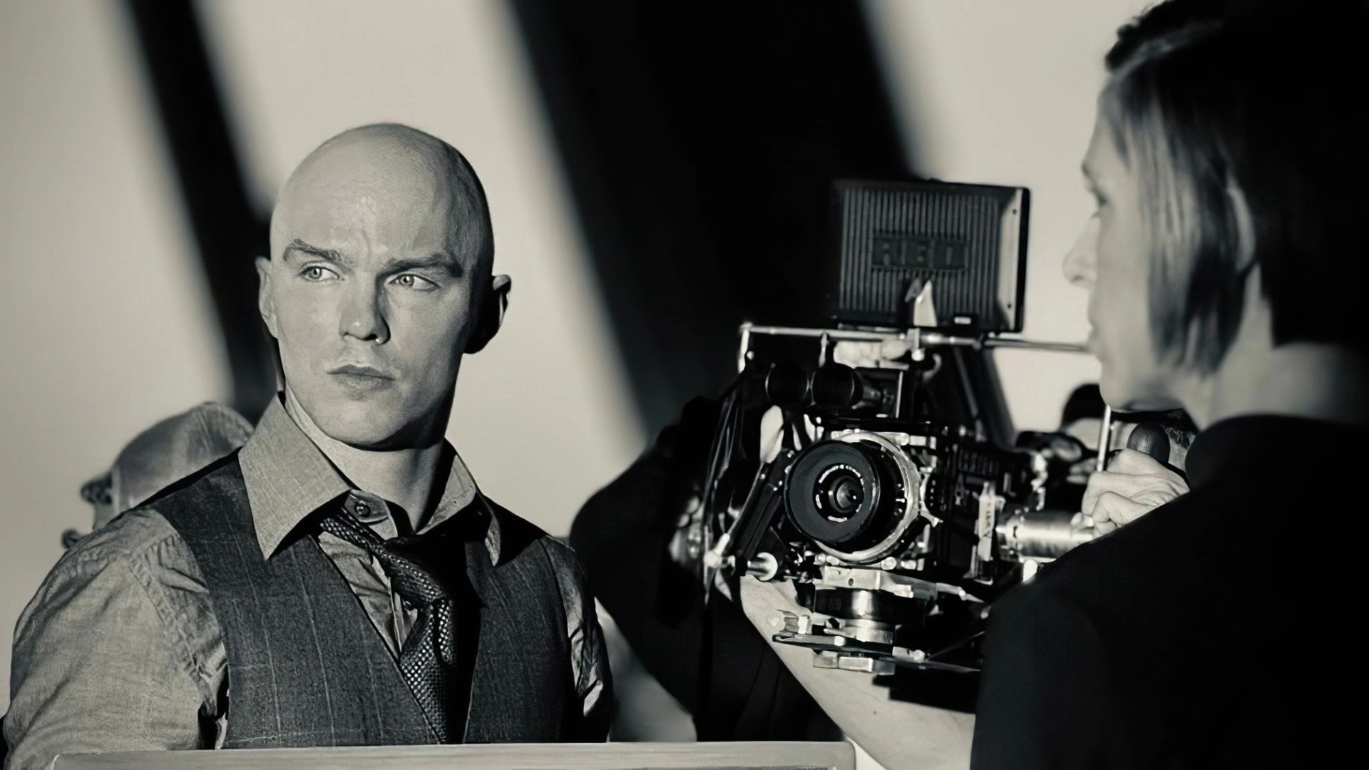 Nicholas Hoult con chaleco y corbata de pie junto a una cámara de cine profesional en un set de rodaje
