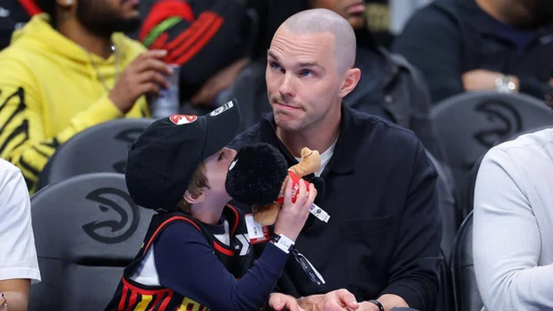 Nicholas Hoult sentado en las gradas de un partido de baloncesto junto a su hijo que sostiene un juguete