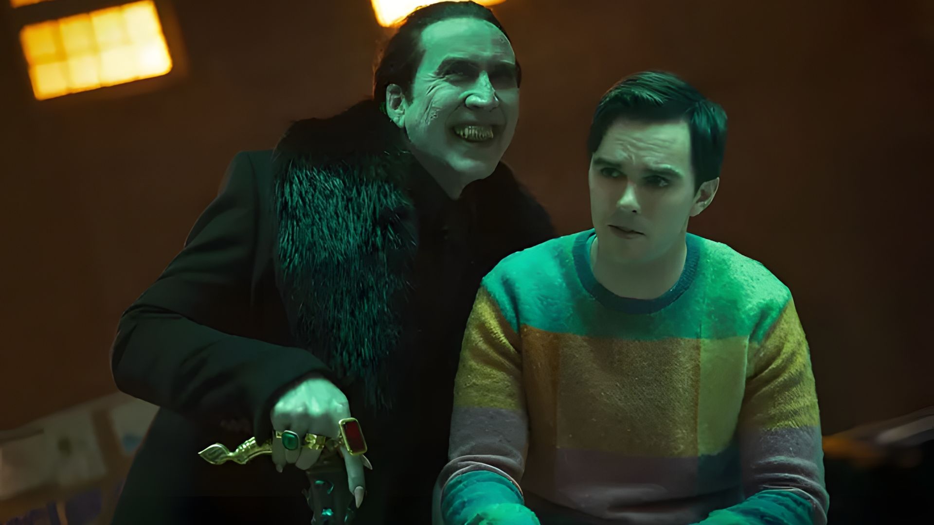 Nicolas Cage con ropa negra y anillos se cierne amenazadoramente sobre Nicholas Hoult con un suéter colorido
