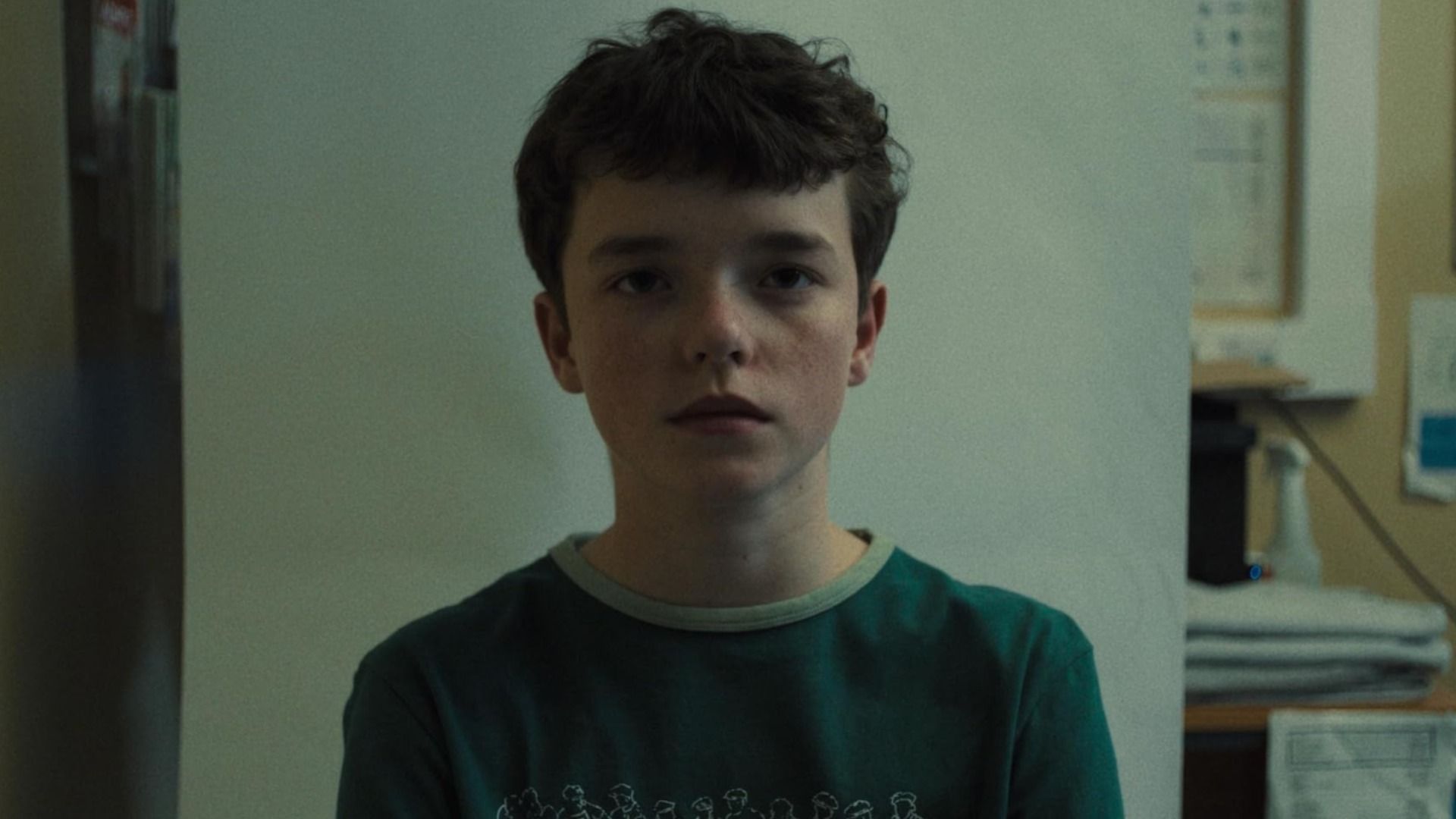 Un chico con cabello oscuro vistiendo una camiseta verde está de pie contra una pared blanca