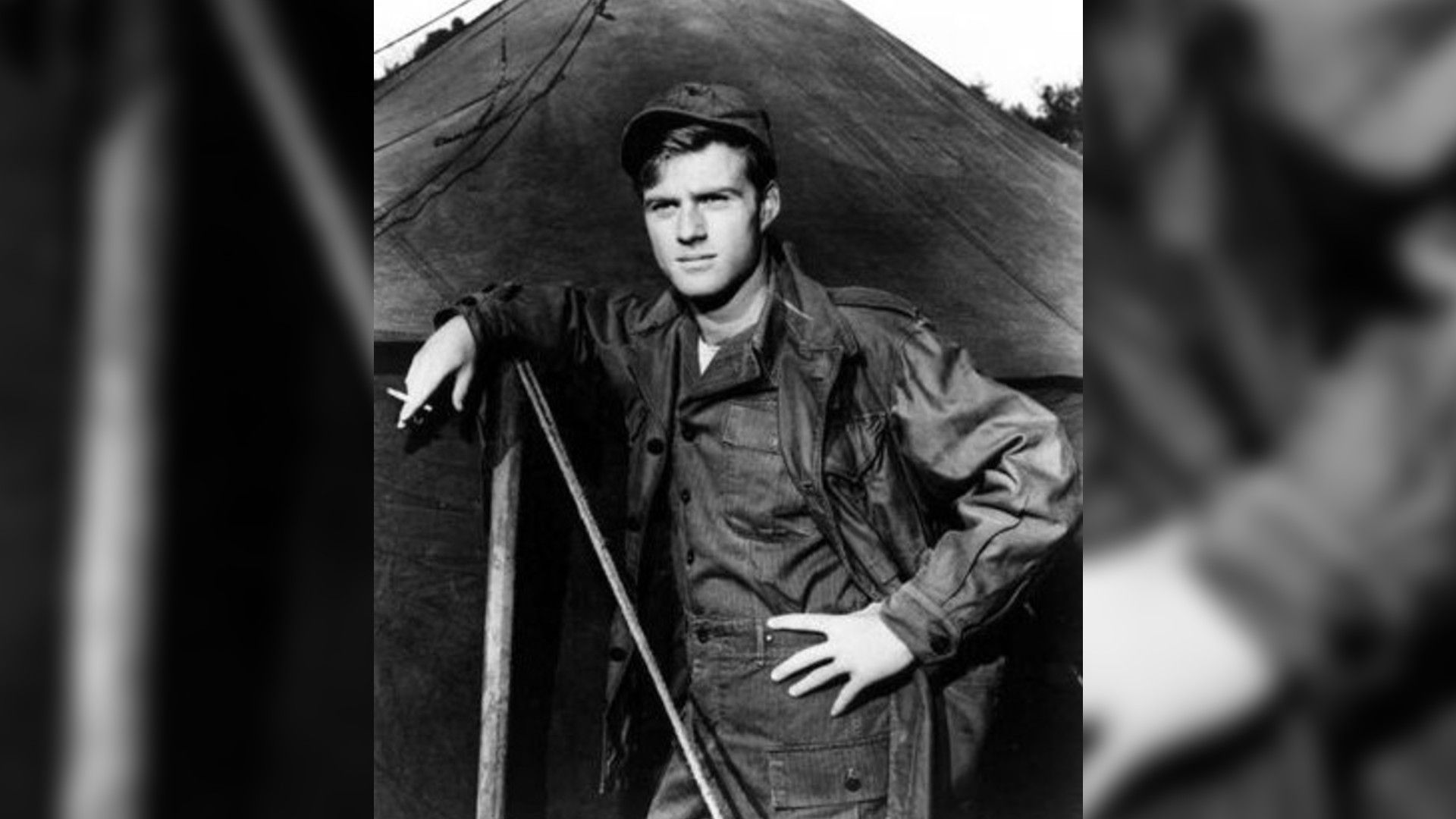 Foto en blanco y negro del joven Robert Redford con uniforme militar de pie junto a una tienda, escena de la película "War Hunt"