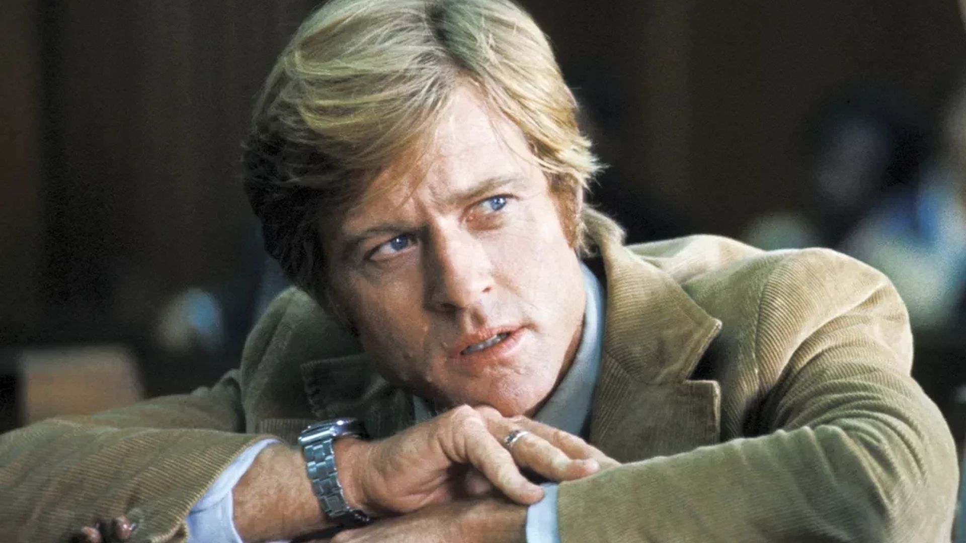 Robert Redford con una chaqueta marrón y un reloj en la muñeca se apoya en las manos y mira hacia un lado