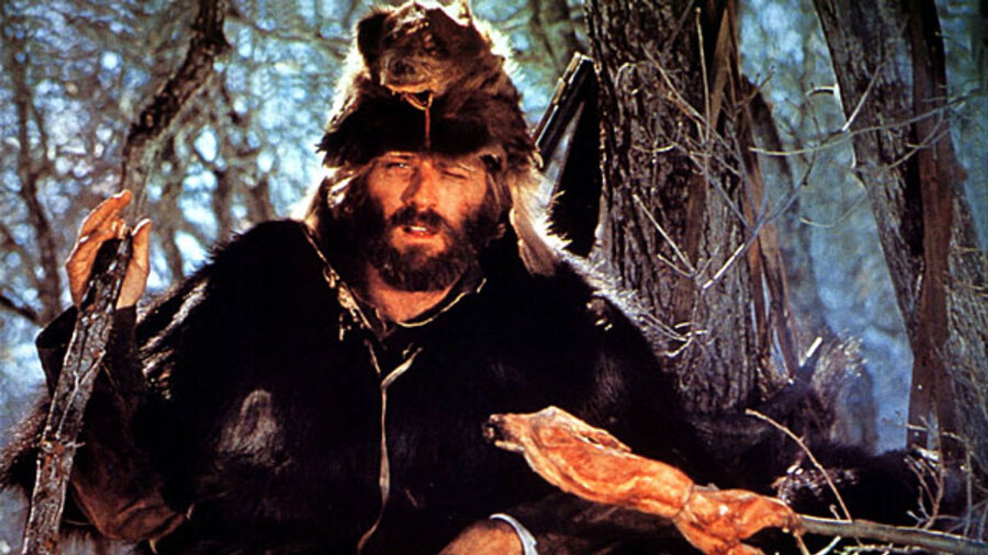Robert Redford con ropa de piel y gorro de cabeza de animal asando carne sobre una hoguera en el bosque, escena de la película "Jeremiah Johnson"
