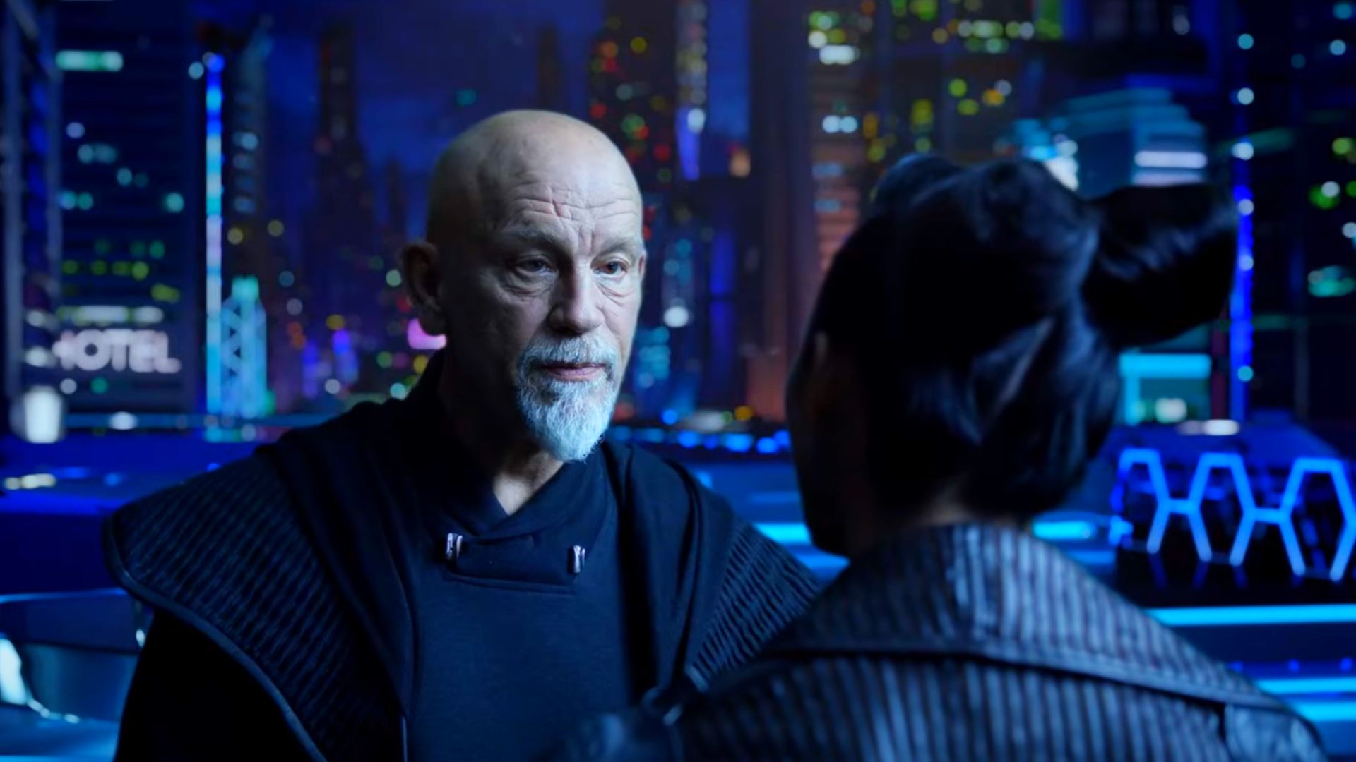 El actor John Malkovich con ropa futurista se encuentra contra el telón de fondo de una ciudad nocturna futurista
