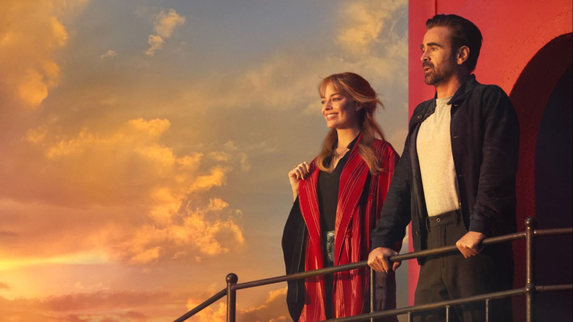 Los actores Margot Robbie y Colin Farrell de pie con ropa casual en la cubierta de un barco viendo la puesta de sol