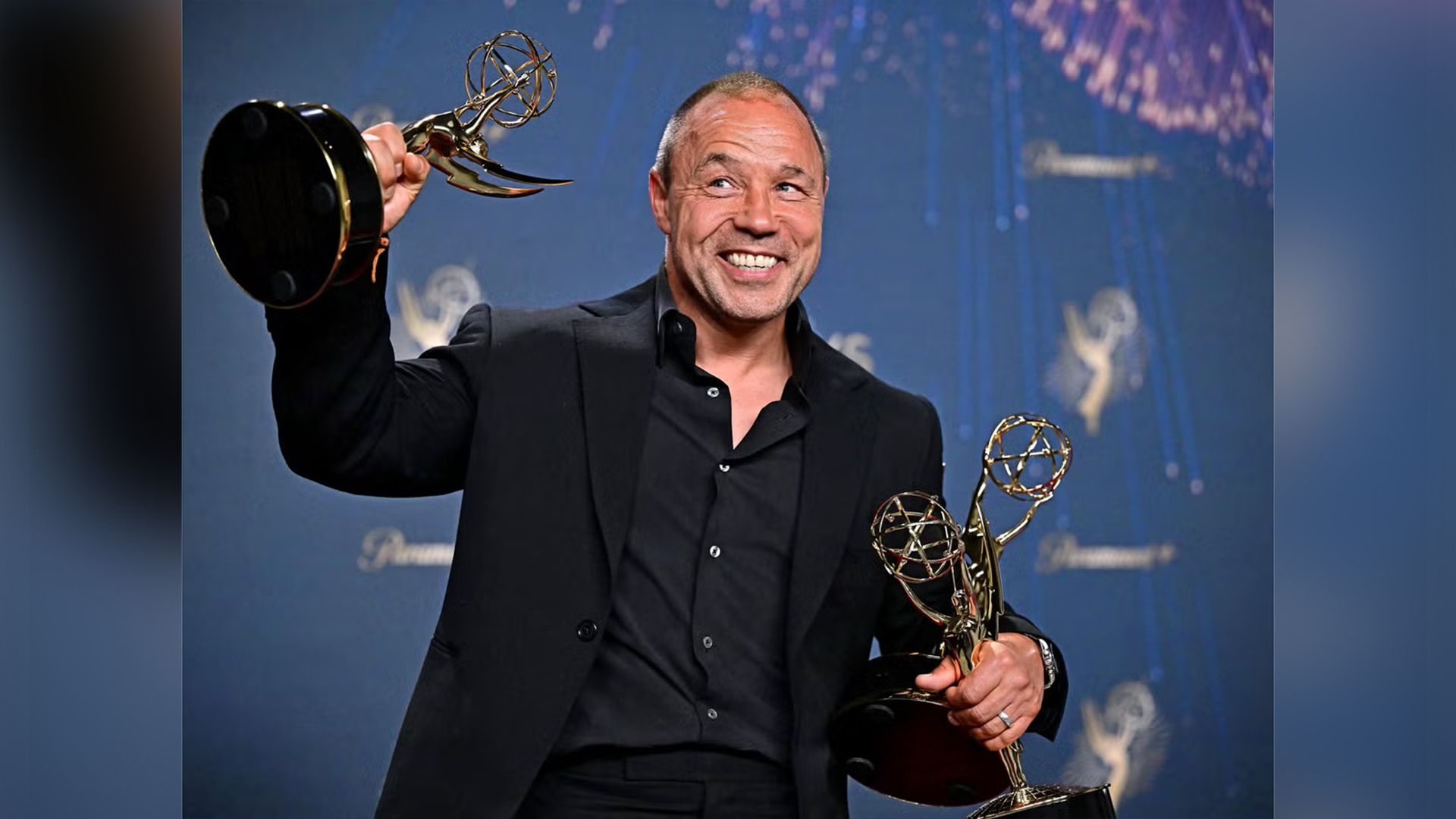 Stephen Graham con traje negro sostiene tres estatuillas doradas de los Emmy en sus manos, sonriendo ampliamente