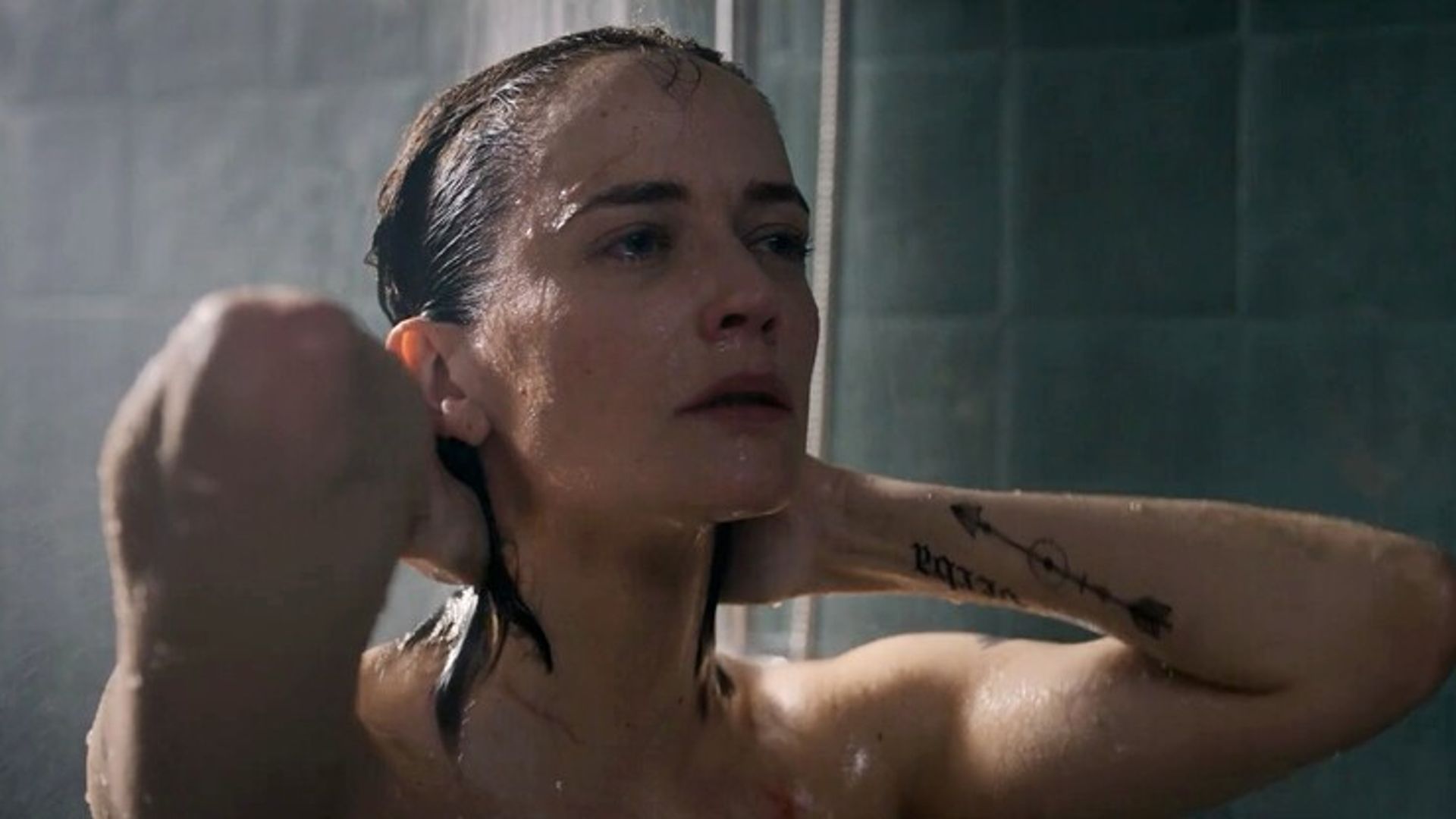 Eva Green con cabello mojado duchándose, manos detrás de la cabeza, solo su rostro y brazo tatuado visibles