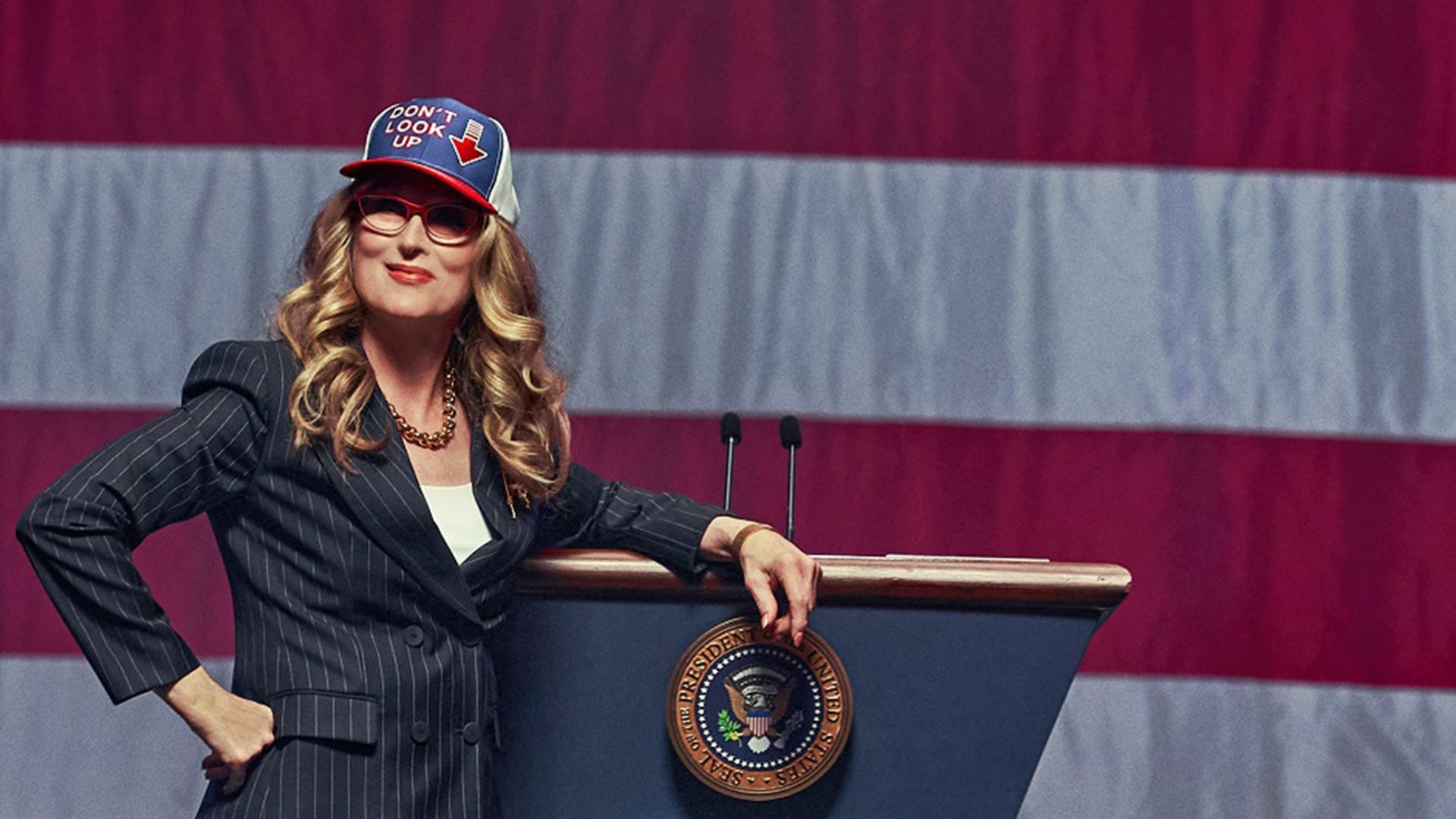Meryl Streep con traje y gorra con la bandera estadounidense se encuentra contra un fondo de la bandera de EE.UU., apoyándose en un podio