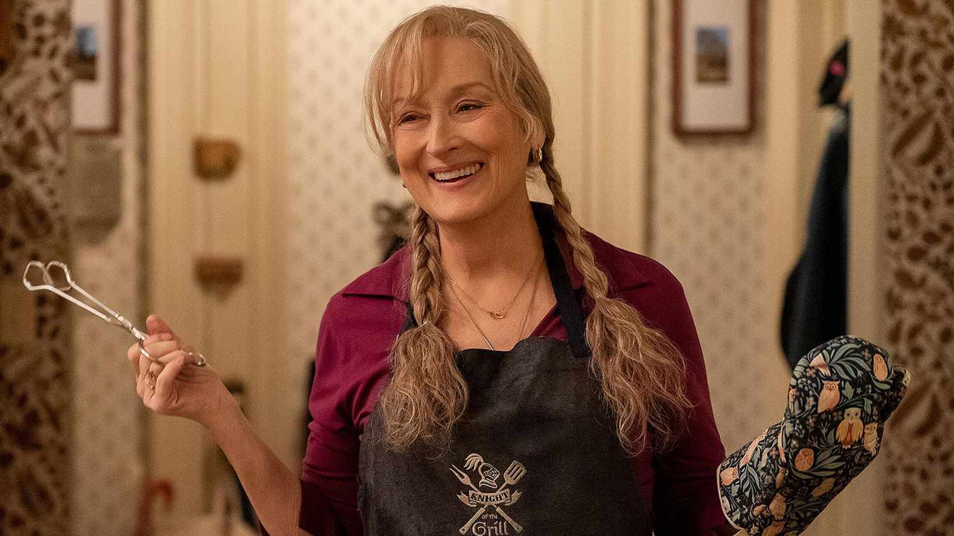 Meryl Streep con trenzas sostiene unas pinzas en una mano, llevando un guante de cocina en la otra