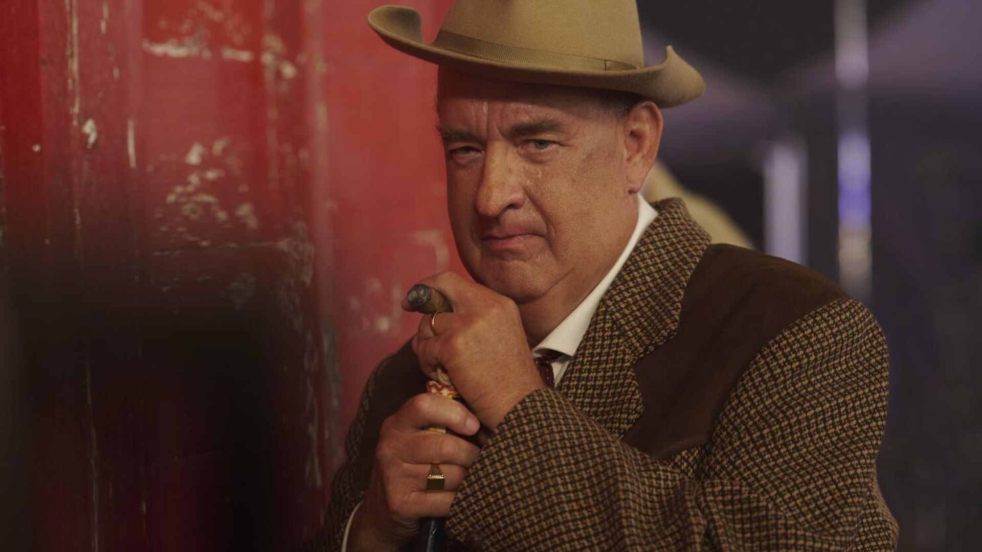 Tom Hanks con traje marrón y sombrero se apoya en un bastón, mirando seriamente hacia la cámara