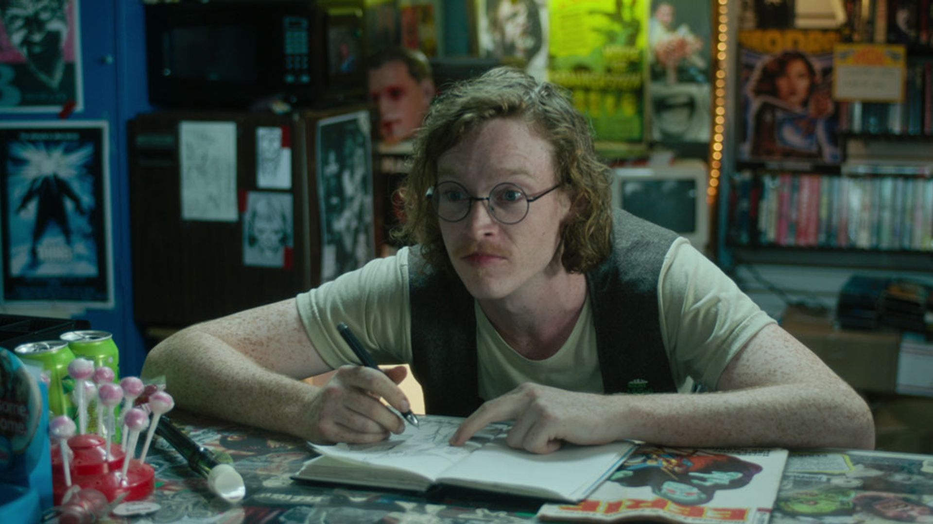 Caleb Landry Jones llevando gafas y un chaleco sentado en una mesa con un bloc de notas, dibujando y mirando hacia un lado