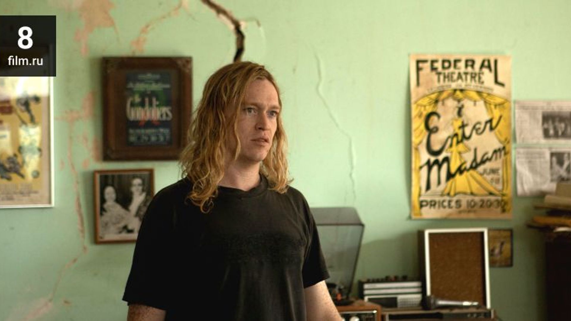Caleb Landry Jones con cabello rubio largo y camiseta negra está de pie en una habitación con pósters en la pared
