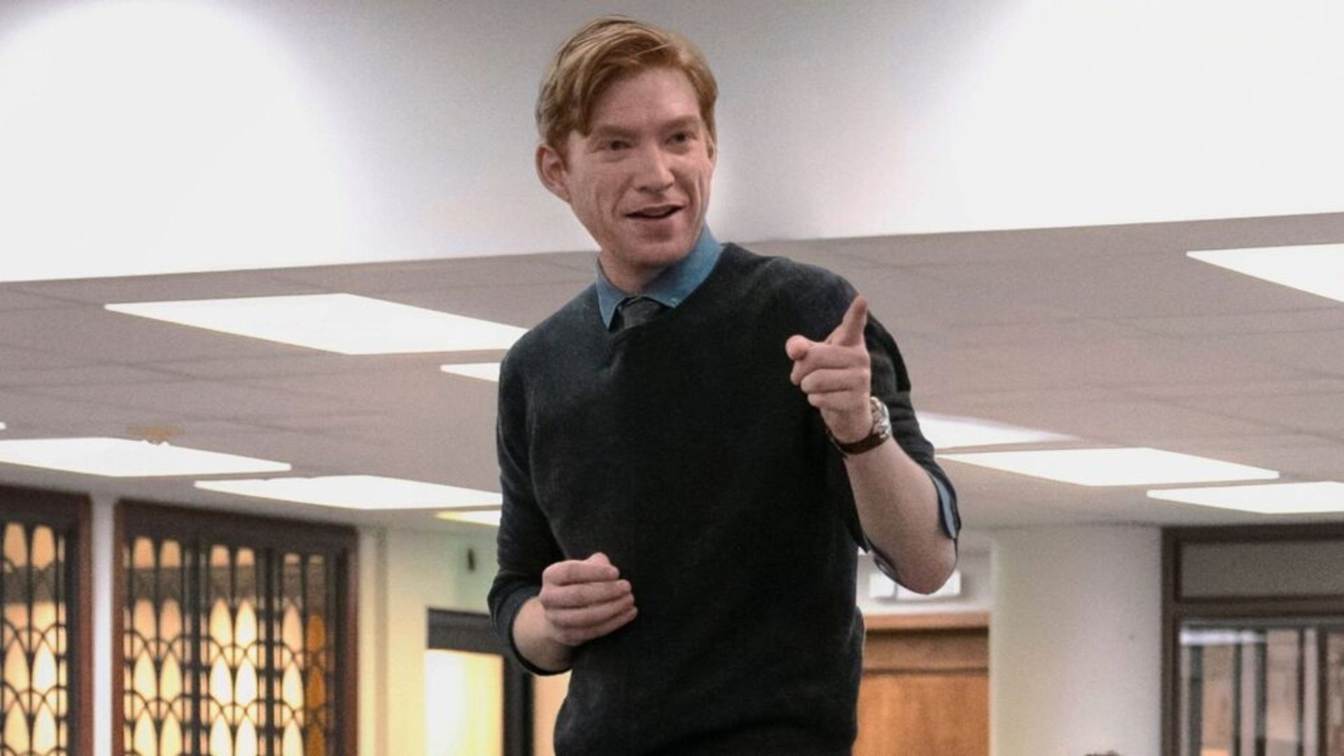 Domhnall Gleeson con una sudadera azul oscuro de pie sobre una mesa y señalando con el dedo