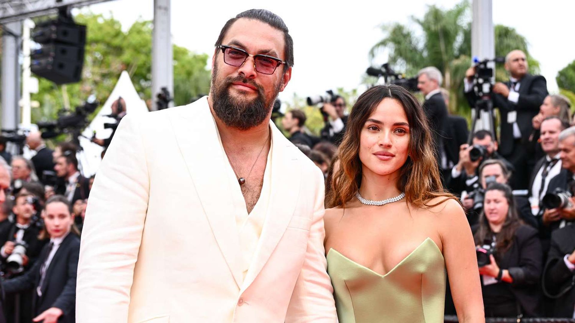 Jason Momoa con traje blanco y Adria Arjona con vestido verde oliva en la alfombra roja