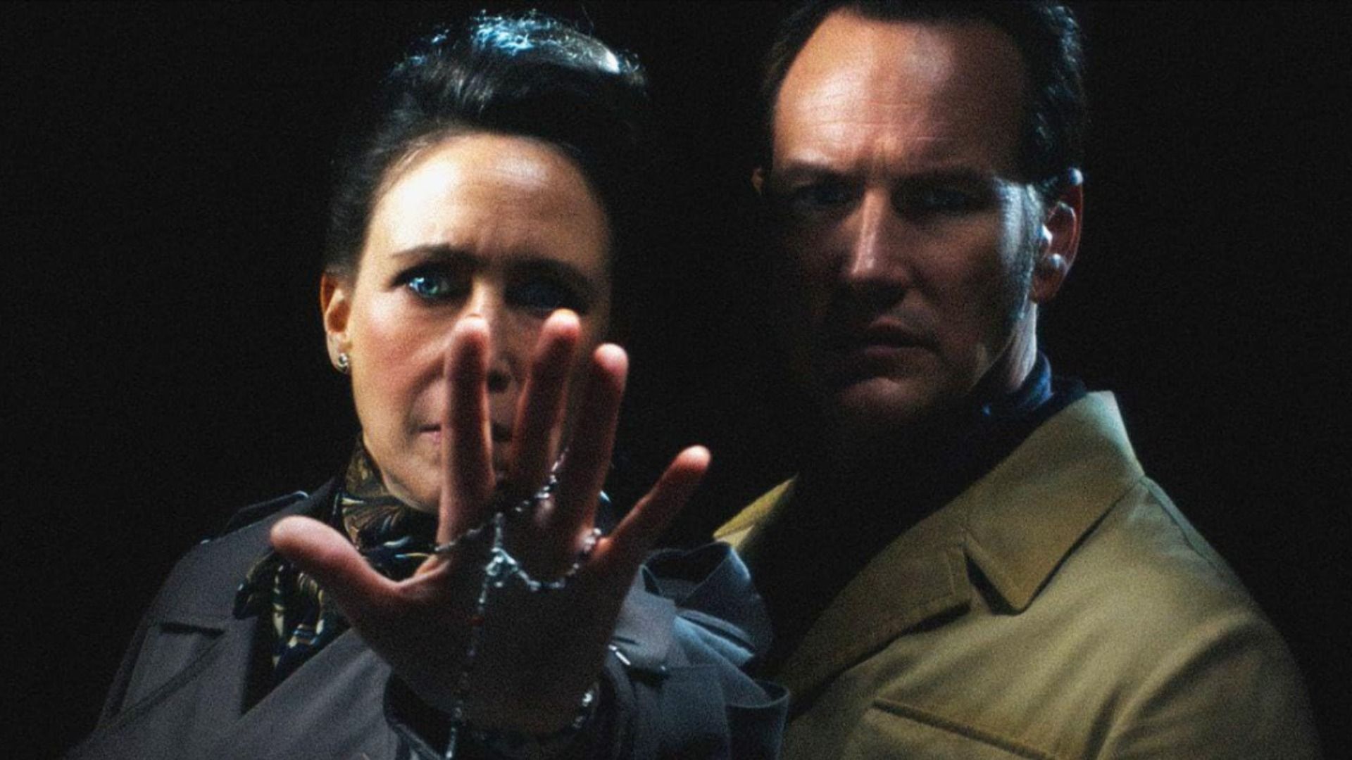 Los actores Vera Farmiga y Patrick Wilson con abrigos de pie contra un fondo negro mirando a la cámara
